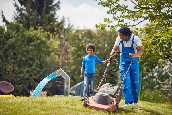 若いアフリカ系アメリカ人の父親は 幼い息子と屋外で草を刈っている 裏庭で一緒に働いている トータル ライフスタイルのコンセプト — ストック写真