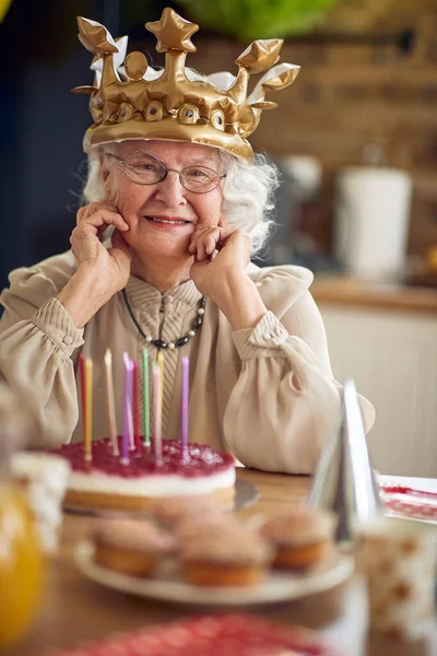 一位慈祥的祖母坐在桌旁 脸上挂着灿烂的微笑 这反映了她一生的爱和智慧 她面前是生日蛋糕 — 图库照片