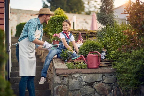 Happy family working outdoors. Young father and girl planting flowers in the flower garden by the house on a sunny summer day, feeling cheerful. Home, family, lifestyle concept.
