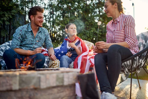 Joyful and happy family of man, woman and girl sitting outdoors by a fireplace roasting marshmallows enjoying sunny summer day together. Family, holiday, lifestyle concept.