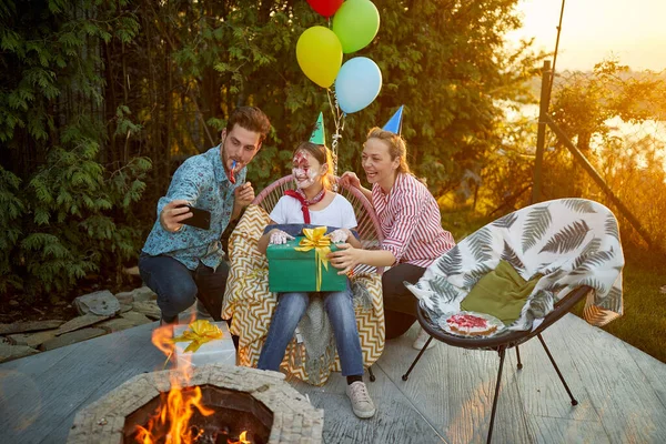 Joyful and festive family of three, celebrating girls birthday, taking a picture outdoors in the backyard by a fireplace. Family, celebration, togetherness concept.
