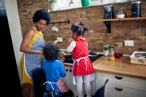 若い母親は ストーブのそばに立っている家庭用キッチンで彼女の2人の幼い子供と夕食を調理し 鍋で野菜を揚げました 楽しさ 家族のコンセプト ストック写真