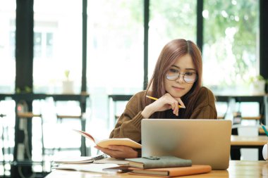 Genç Asyalı gözlüklü kız öğrenci online kuzenlerini öğreniyor ve masa başında oturup dizüstü bilgisayar ve ders kitapları kullanarak notlar yazıyor. Çevrimiçi çalışma ve öğrenme kavramı.