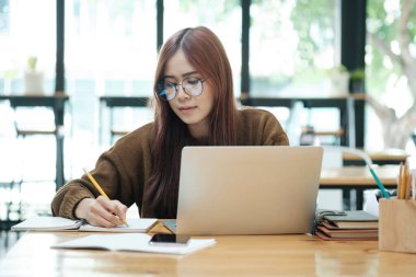 Genç Asyalı gözlüklü kız öğrenci online kuzenlerini öğreniyor ve masa başında oturup dizüstü bilgisayar ve ders kitapları kullanarak notlar yazıyor. Çevrimiçi çalışma ve öğrenme kavramı.