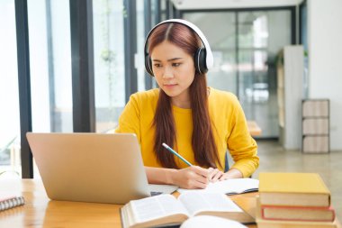 Genç Asyalı gözlüklü kız öğrenci online kuzenlerini öğreniyor ve masada otururken notlar yazıyor ve dizüstü bilgisayar ve kulaklık takıyor. Çevrimiçi çalışma ve öğrenme kavramı