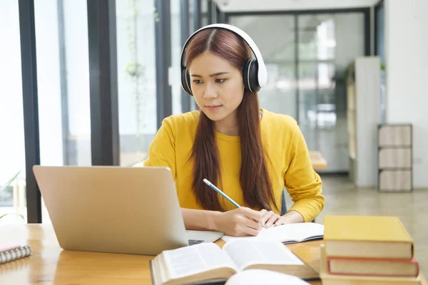 若いアジアの女性学生は オンラインコースを学び 机に座りながらノートを書き ヘッドフォンを装着したラップトップや教科書を使用して眼鏡をかけています オンライン学習と学習の概念 — ストック写真
