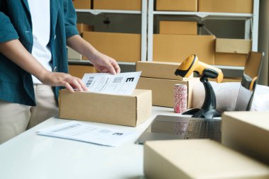 Küçük işletme girişimcisi genç bir kadın, arka planında karton kutu rafları olan bir mağaza sahibi posta kutusunda ürünleri paketliyor, müşteriye sevkiyat için teslimat paketini etiketliyor. Çevrimiçi