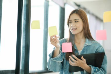 Genç kız öğrenci, elinde tablet tutarken cam duvara yapışmış post-it 'lerle çalışma planları ya da yaratıcı proje fikirleri öğreniyor. Çevrimiçi öğrenme kavramı.