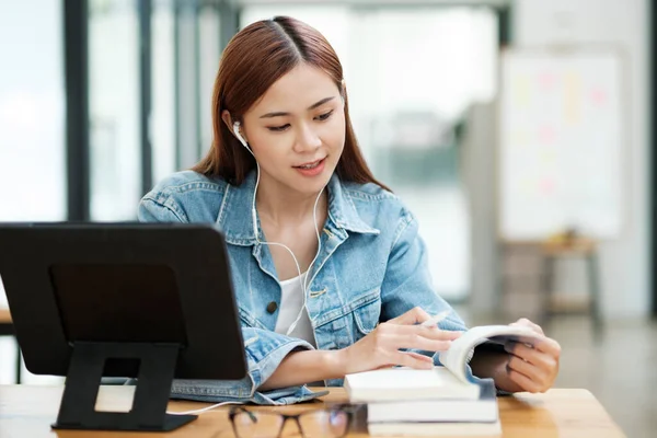 Young asian woman studying online using tablet and smart pen looking at books wearing earphones while sitting at desk with textbooks and glasses at indoor room. Online learning concept.