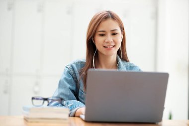 Genç Asyalı kız öğrenci dizüstü bilgisayar kullanarak ve kulaklık takarak çalışırken gözlüklü ve ders kitaplarıyla internetten eğleniyor. Çevrimiçi öğrenme kavramı.