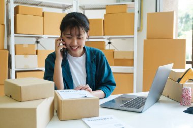 Genç bayan küçük işletme girişimcisi, dizüstü bilgisayarda çalışan bir dükkan sahibi depoda kutuları olan müşteriyle telefon görüşmeleri yapan, siparişi doğrulayan. Çevrimiçi satış, e-ticaret kavramı.