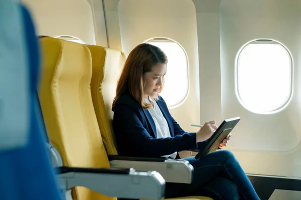 窓の近くの飛行機のキャビンに座っている間 スマートペン書き込みとタブレットを使用して作業正式な服で若いプロのアジアのビジネスマンの女性は別の場所に移動します 旅とビジネスのコンセプト — ストック写真