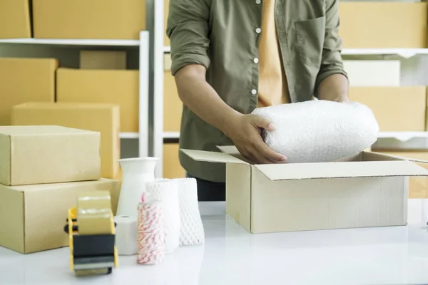 Young small business entrepreneur man, online store owner with shelves of boxes in the background packing product in mailing box at workplace for shipping to customer. selling product online. Online shopping concept.