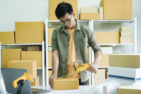 Young small business entrepreneur man, online store owner with shelves of boxes in the background packing product in mailing box sealing box with tape at workplace for shipping to customer. Online shopping concept.