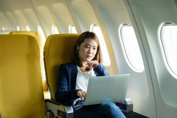 stock image Young professional asian businesswoman in formal clothes working using laptop on her lap while sitting in airplane cabin near window traveling to another place. Traveling and business concept.