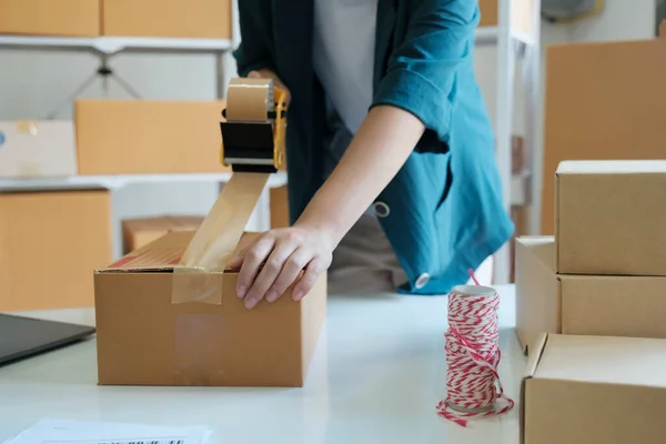 Young small business entrepreneur woman, online store owner with shelves of boxes in the background packing product in mailing box sealing box with tape at workplace for shipping to customer. Online shopping concept.