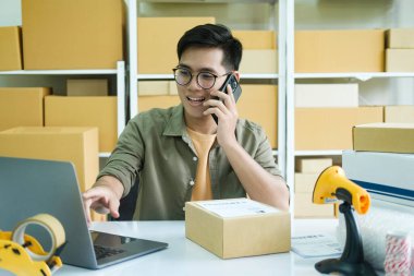 Genç erkek, küçük işletme girişimcisi, dizüstü bilgisayarda çalışan, depo masasında kutularla çalışan müşteriyle telefon görüşmeleri yapan, siparişi doğrulayan. Çevrimiçi satış, e-ticaret kavramı..