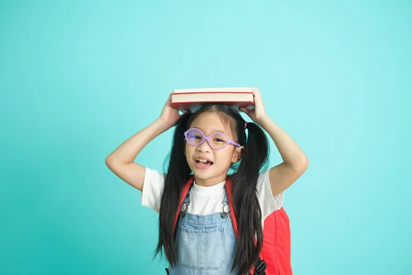 stock image Portrait of happy lazy little girl funny smiling, cute, child school girl with glasses hold books on her head while in the classroom, disobedient child having fun.indoor studio shot isolated on blue background.Educational concept for school.