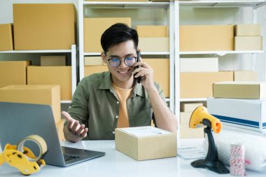 Genç erkek, küçük işletme girişimcisi, dizüstü bilgisayarda çalışan, depo masasında kutularla çalışan müşteriyle telefon görüşmeleri yapan, siparişi doğrulayan. Çevrimiçi satış, e-ticaret kavramı..