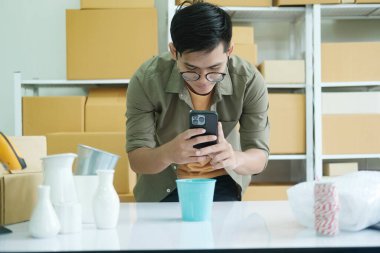 Asyalı erkek, çevrimiçi işyeri sahibi veya çevrimiçi dükkan sahibi cep telefonu kullanıyor fotoğraf çekiyor ve paket kutusunu kontrol ediyor, teslimat için hazırlanan kargo kutusu üzerindeki perakende paket barkodunu tarıyor ve...