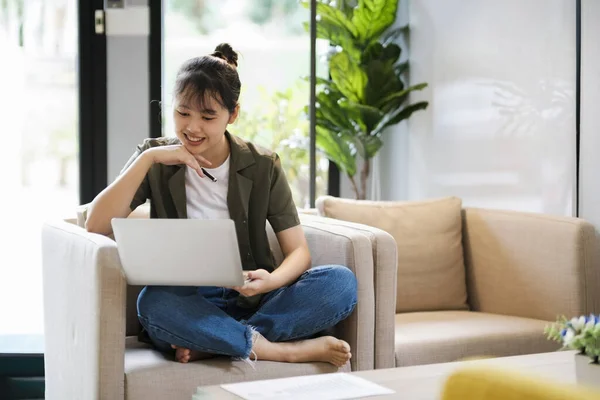 年轻的亚洲女人在家里坐在沙发上 一边学习 一边上网 一边用耳机听音乐 收听播客 有声书籍 使用笔记本电脑观看录像 — 图库照片