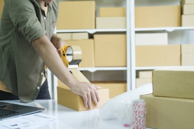 Küçük işletme girişimcisi, çevrimiçi mağaza sahibi, arka planda raflar dolusu kutuları olan, posta kutusundaki kutuları paketleyen, iş yerindeki müşteriye gönderilmesi için kaset taşıyan bir adam. Çevrimiçi alışveriş kavramı.