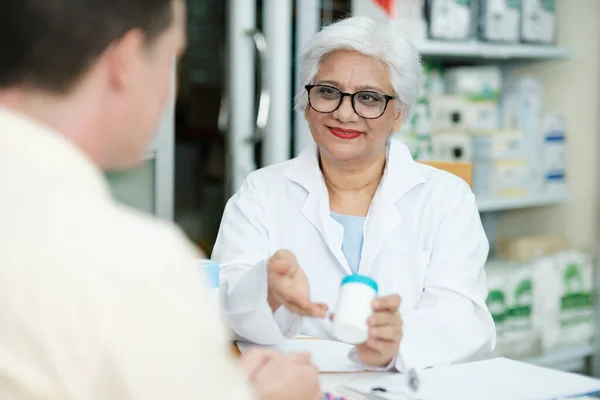 Female senior pharmacist at the drugstore wearing white gown talking, giving advice, explaining, suggesting, and recommending to client or patient about the prescription and medications. Medicine and
