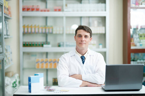 Male smart young-adult pharmacist in professional white gown working at desk in modern pharmacy, drugstore using laptop to check or record information. Medicine and healthcare concept.