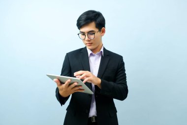 Gözlük takan ve gülümseyen genç Asyalı iş adamının portresi. Tableti çalışırken ya da açık mavi izole arka planla iletişim kurarken kullanıyor. İş, bağlantı kavramı.