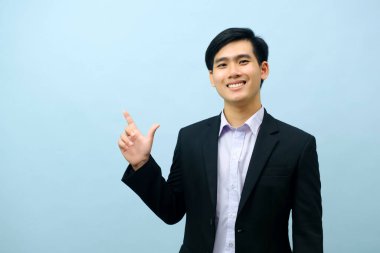 Portriat of asian young smart happy businessman dressed in suit standing straight, smiling, and pointing finger looking at camera with isolated light blue background. Business concept.