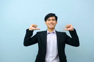 Portriat of asian young smart happy businessman dressed in suit standing straight, smiling, and pointing thumb finger at himself looking at camera with isolated light blue background. Business concept