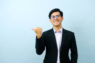 Portriat of asian young smart happy businessman dressed in suit standing straight, smiling, and pointing finger looking at camera with isolated light blue background. Business concept.