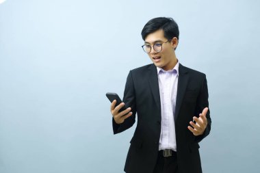 Portrait of a surprised young smart asian businessman looking at his smart phone screen in disbelief with light blue background. Concept of business.