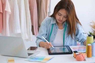 Young asian tailor designer designing on a new fashion collection by using tablet. She is sketching and drawing on a digital tablet at the table in her design studio.