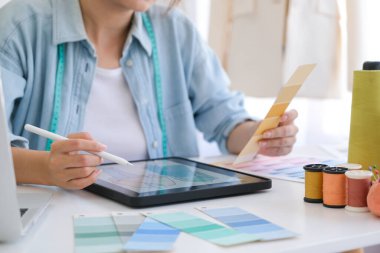 Young asian tailor designer designing on a new fashion collection by using tablet. She is sketching and drawing on a digital tablet at the table in her design studio.