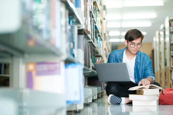 若い男性の大学生 眼鏡をかけた高校生 読書本の上に座っているカジュアルな服を着て 図書館で学校プロジェクトのためのラップトップを使用して研究を勉強し 研究をしています Learning Educationの概念 — ストック写真