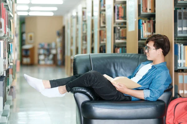 本を読み 図書館で学校プロジェクトのために勉強し 研究を行うソファに座って眼鏡やカジュアルな服を着て若い男性の大学生 Learning Educationの概念 — ストック写真