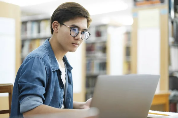 眼鏡をかけた若い男性大学生や 机に座ってノートパソコンを使って本を勉強したり 図書館で本を読んだりして 研究や学校プロジェクトの情報を探したりしています Learning Educationの概念 — ストック写真