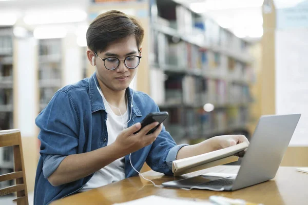 年轻的男大学生戴着眼镜 头戴耳机 身穿休闲装 坐在课桌前 用笔记本电脑和手机看书 研究或实施学校项目 — 图库照片