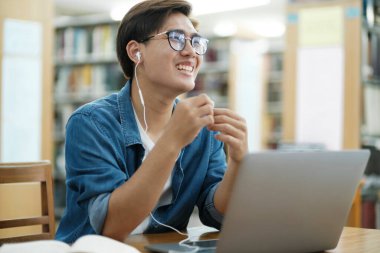 Genç erkek üniversite öğrencisi gözlük takıyor ve gündelik kıyafetlerle masa başında oturmuş, çalışmak ve kitap okumak için kulaklık takıyor ve kütüphanede araştırma ya da okul projesi için müzik dinliyor. Eğitim ve E-Öğrenme kavramı.