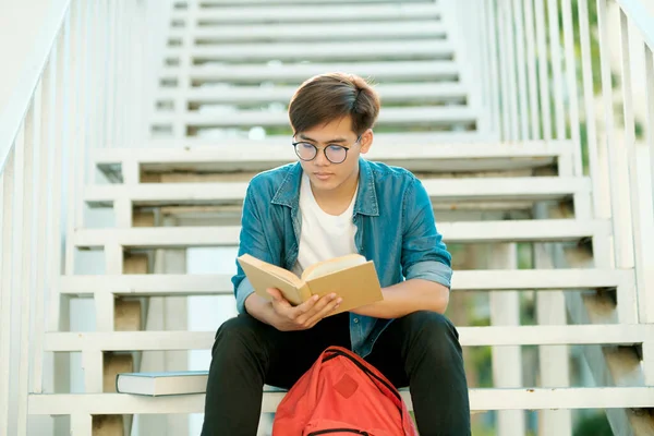 Young male college student with eyeglasses and in casual clothings sitting outdoor on stairs to study and read books alone looking for information for school project and research. Education concept.