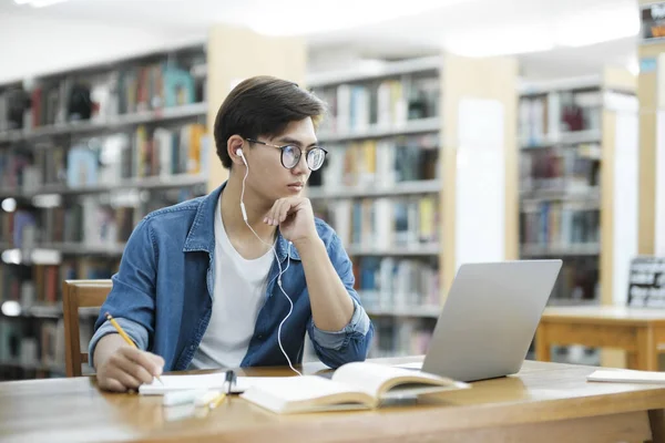 年轻的男大学生戴着眼镜 身穿休闲装 坐在课桌前学习 认真思考 并在图书馆或学校项目中佩戴耳机 用笔记本电脑写下便条 学习概念 — 图库照片
