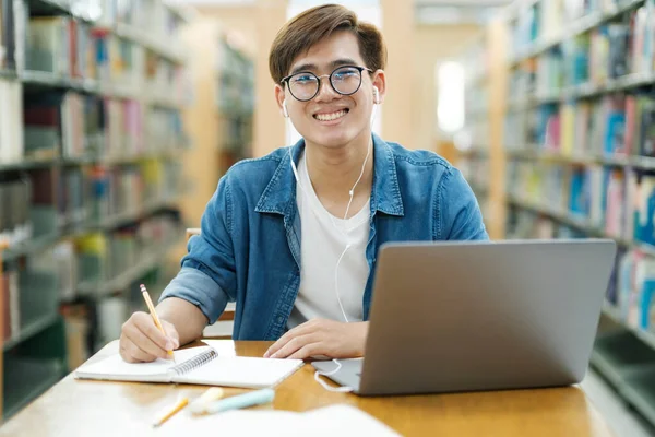 年轻的男大学生戴着眼镜 身穿休闲装 坐在课桌前学习 用手提电脑记笔记 带着耳机在图书馆观看研究或学校项目 学习概念 — 图库照片