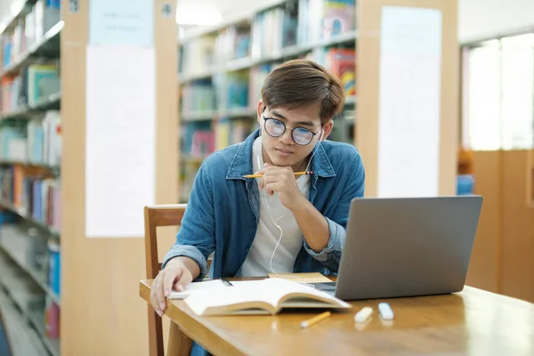 年轻的男大学生戴着眼镜 身穿休闲装 坐在课桌前学习 用笔记本电脑写笔记 带着耳机参加研究或学校项目 电子学习概念 — 图库照片
