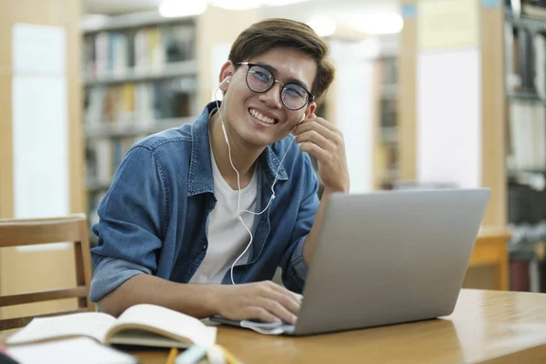 年轻的男大学生戴着眼镜 身穿休闲装 坐在课桌旁 用笔记本电脑 头戴耳机 阅读书籍 并在图书馆听音乐 准备参加研究或学校项目 教育和电子学习概念 — 图库照片