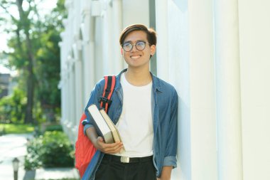 Gözlüklü, günlük kıyafetleri olan ve arka planda kampüs binası olan sırt çantalı genç ve zeki erkek kolaj öğrencisi. Çalışmaya hazır kameralara bakarken kitapları tutuyor. Eğitim kavramı.