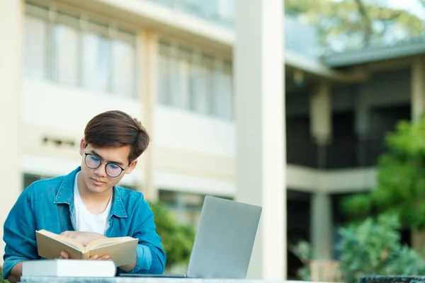 年轻的男大学生 戴着眼镜 身穿休闲装 坐在室外学习 读书和看书 他们使用笔记本电脑搜寻学校项目和研究的信息 教育和电子学习概念 — 图库照片