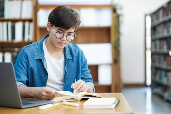 眼鏡をかけ 学内プロジェクトや試験準備のために図書館の机の上にノートパソコンを使ってノートを勉強したり 読んだり 書き留めたりする若い男性大学生 Learning Educationの概念 — ストック写真