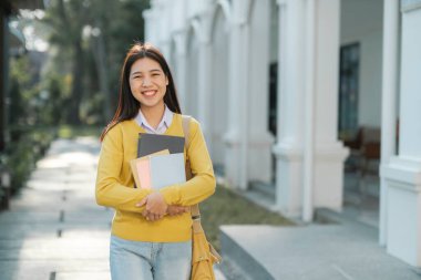 Sırt çantasıyla sırtüstü gülümseyen, arka planda kampüs binası olan ve çalışmaya hazır kameralara bakarken elinde kitaplarla bekleyen zeki kız kolaj öğrencisi. Eğitim kavramı.