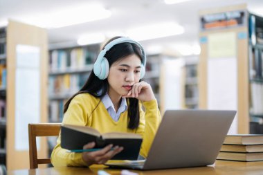 Genç bayan kolej, kulaklık takan lise öğrencisi masa başında oturmuş kitap okuyor ve kütüphanedeki okul projesi için dizüstü bilgisayarla araştırma yapıyor. E-Öğrenme ve Eğitim Konsepti.